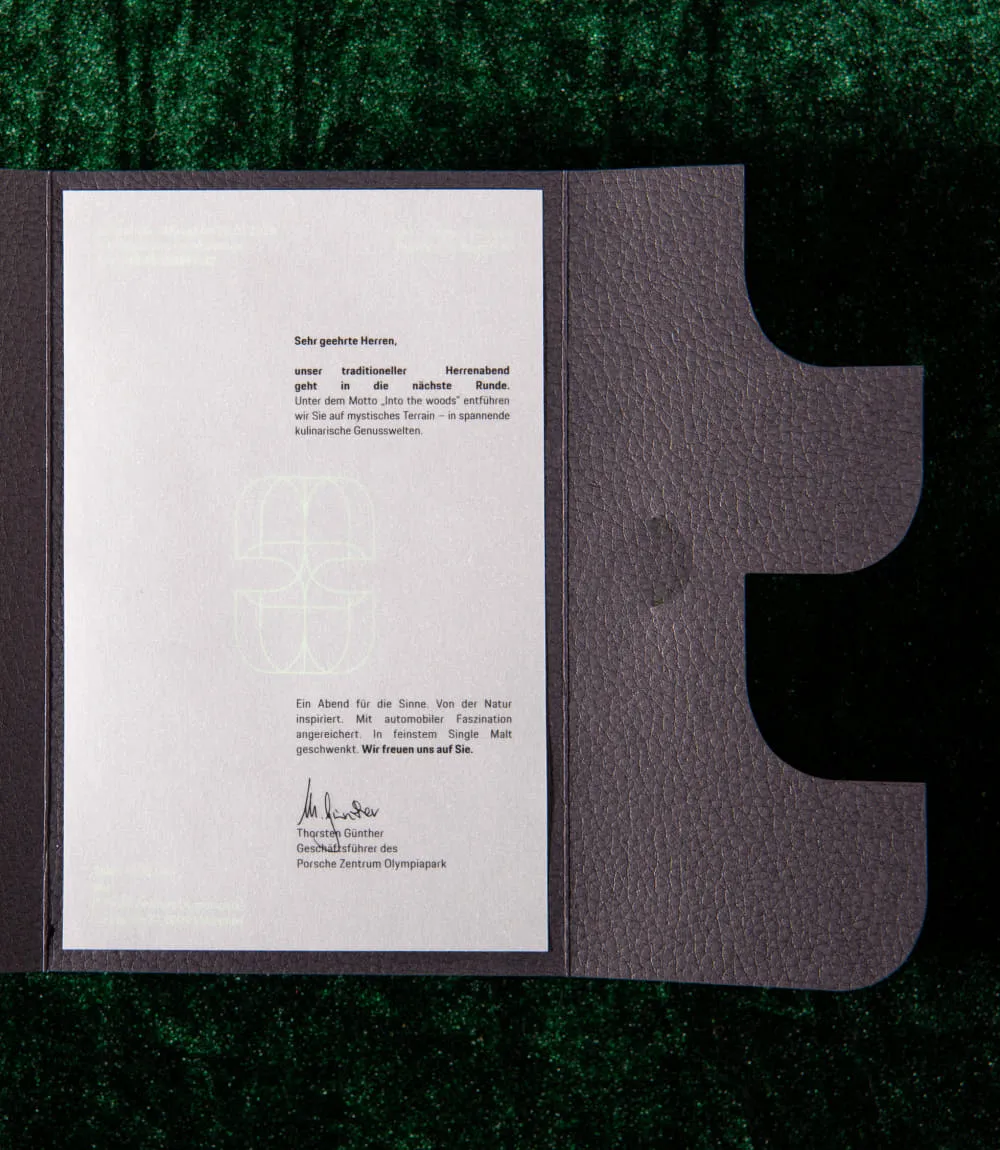 Geöffnete schwarze Mappe in Lederoptik auf dunkelgrünem Satinstoff. Eingelegt ist ein weißes Blatt mit einem Einladungsschreiben zu einem Herrenabend unter dem Motto Into the Woods.