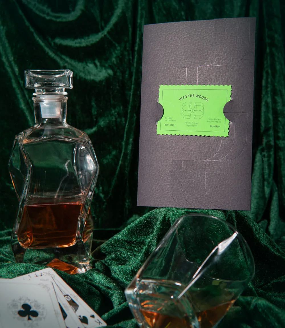 Auf und vor dunkelgrünem Satinstoff drapiert sind rechts eine schwarze Mappe in Lederoptik mit einem grünen Into the Woods Ticket mittig. Links eine halb gefüllte Whisky Flasche und vor der Mappe liegend ein Whiskyglas und drei aufgedeckte Spielkarten. 