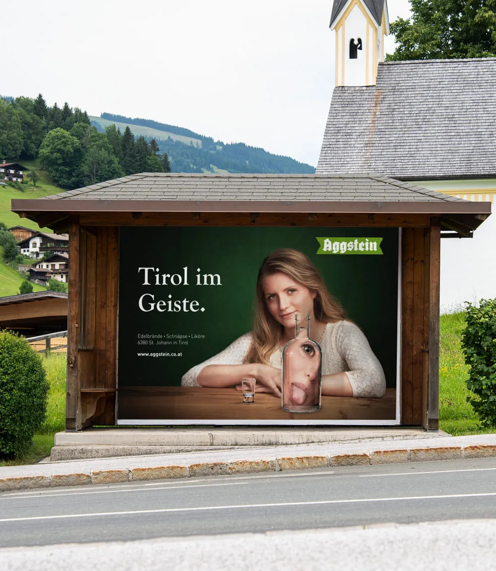 In einem Bushaltestellenhäuschen ist ein großformatiges Werbeplakat von Aggstein zu sehen. Darauf eine junge Frau mit blonden Haaren sitzt an einem Holztisch vor einer dunkelgrünen Wand. Ihr Gesicht wird in einer vor ihr stehenden Glasflasche mit rausgestreckter Zunge verzerrt dargestellt. Daneben steht noch ein passendes Schnapsglas.Links eine Schriftblock in weiß betitelt mit Tirol im Geiste.